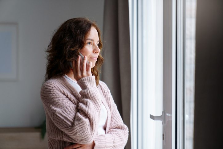 Young woman talking on mobile phone at home, looking through window. Tensed girl holding cell phone and calling, waiting. People connection, communication. Lifestyle moment.